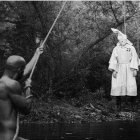 AN IMAGE OF WHAT APPEARS TO BE A BLACK MAN HANGING A WHITE KLANSMAN IN THE WOODS.